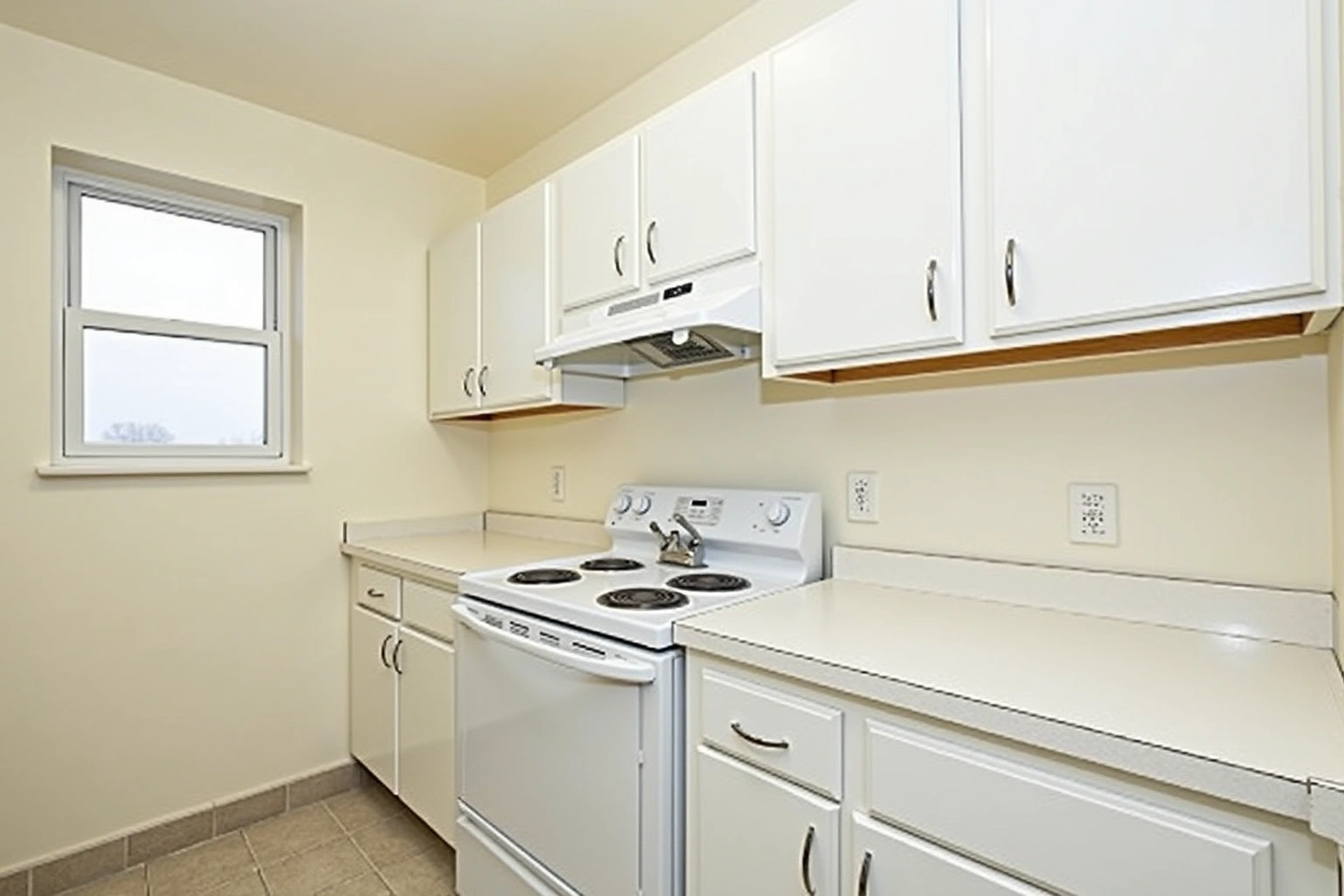 Simple apartment kitchen with white recessed-panel upper and lower cabinets, chrome C-pulls, white laminate counter, a white electric coil range and a single window.